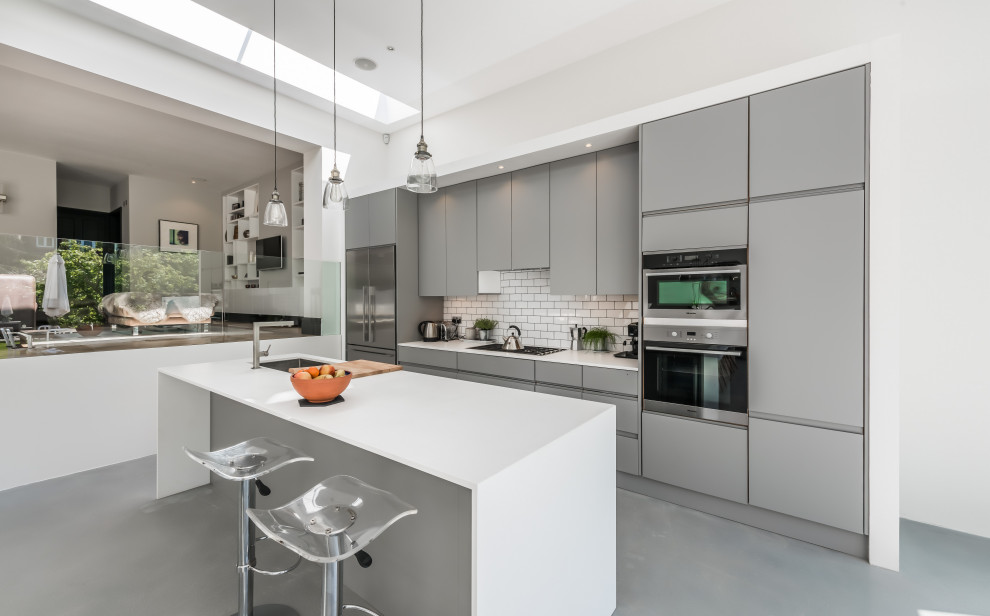 This is an example of a contemporary galley kitchen in London with a submerged sink, flat-panel cabinets, grey cabinets, white splashback, metro tiled splashback, stainless steel appliances, concrete flooring, an island, grey floors and white worktops.