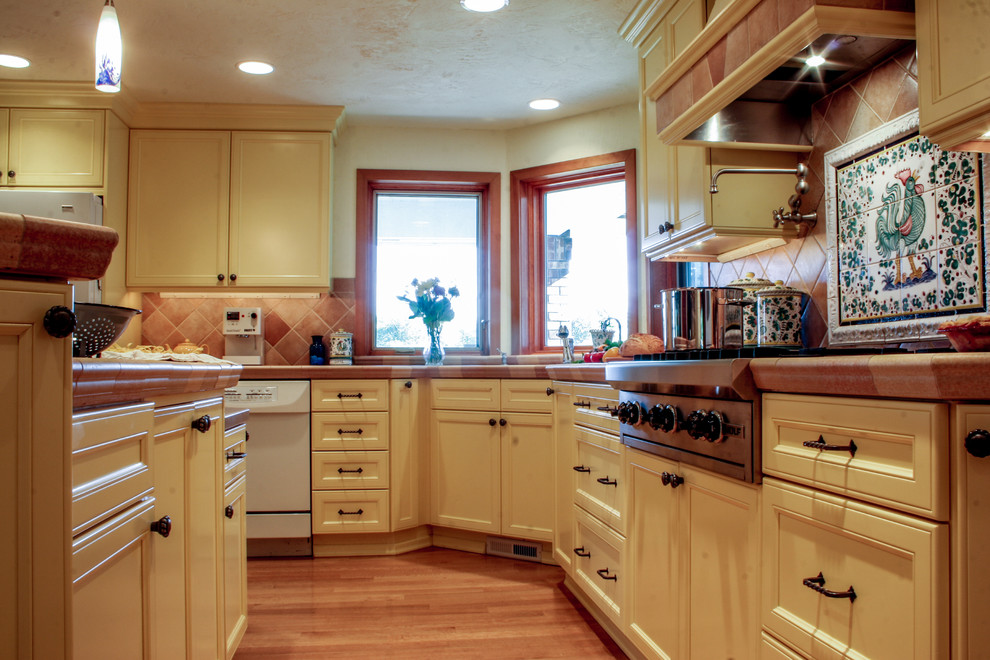 This is an example of a large mediterranean l-shaped kitchen/diner in Seattle with recessed-panel cabinets, yellow cabinets, tile countertops, orange splashback, ceramic splashback, an island, white appliances and light hardwood flooring.