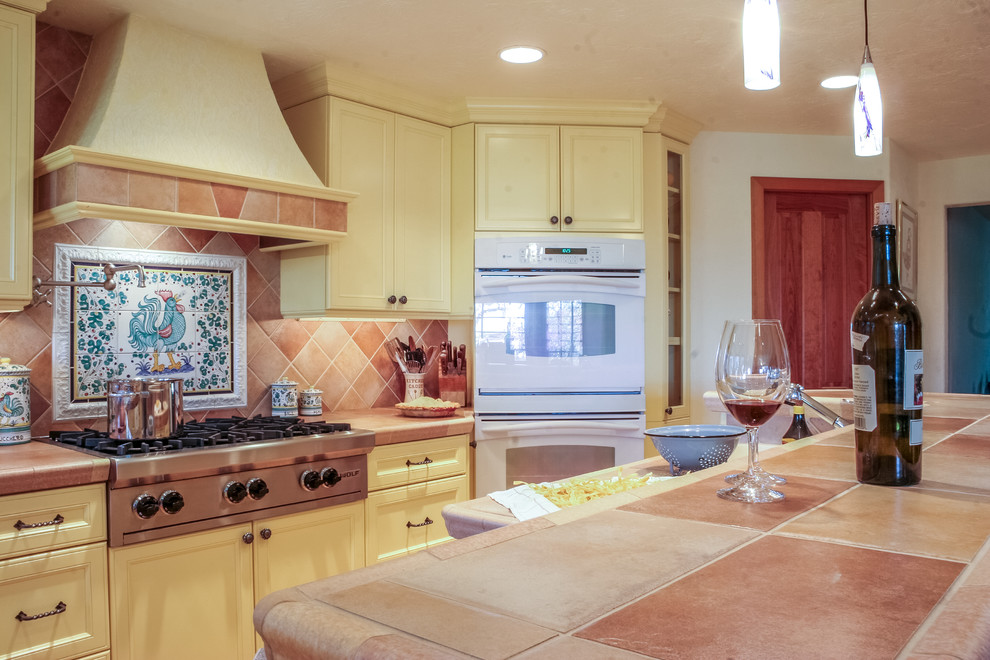 Photo of a large mediterranean l-shaped kitchen/diner in Seattle with a built-in sink, recessed-panel cabinets, yellow cabinets, tile countertops, orange splashback, ceramic splashback, white appliances, light hardwood flooring and an island.