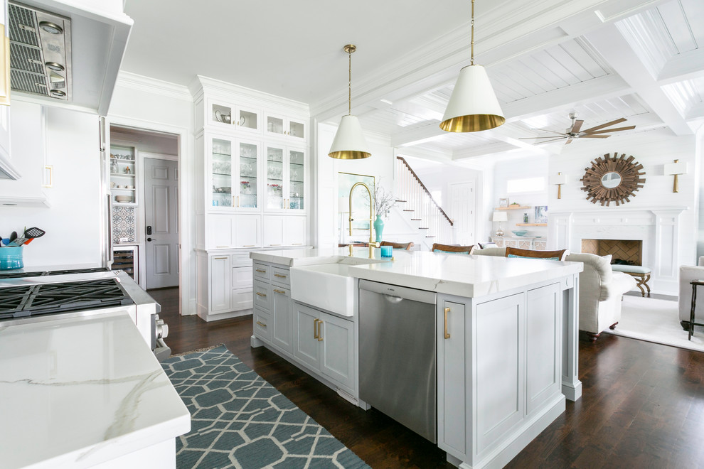 This is an example of a beach style l-shaped kitchen in Charleston with a belfast sink, shaker cabinets, white cabinets, stainless steel appliances, dark hardwood flooring, an island, brown floors and white worktops.