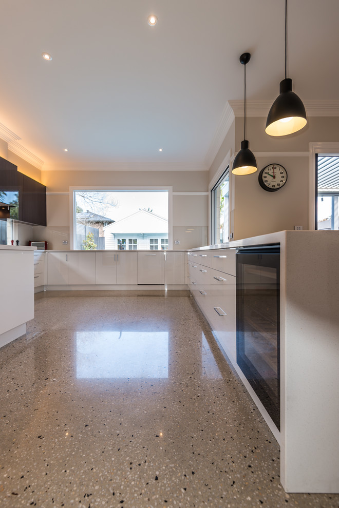 This is an example of a large contemporary open plan kitchen in Melbourne with flat-panel cabinets, white splashback, glass sheet splashback, black appliances, concrete flooring, a built-in sink, white cabinets, engineered stone countertops, grey floors and white worktops.
