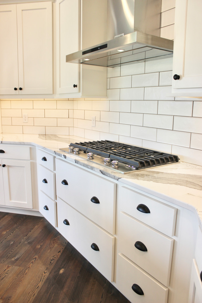 Inredning av ett lantligt stort vit vitt kök, med en rustik diskho, släta luckor, vita skåp, bänkskiva i kvarts, vitt stänkskydd, stänkskydd i tunnelbanekakel, rostfria vitvaror, mellanmörkt trägolv, en köksö och brunt golv