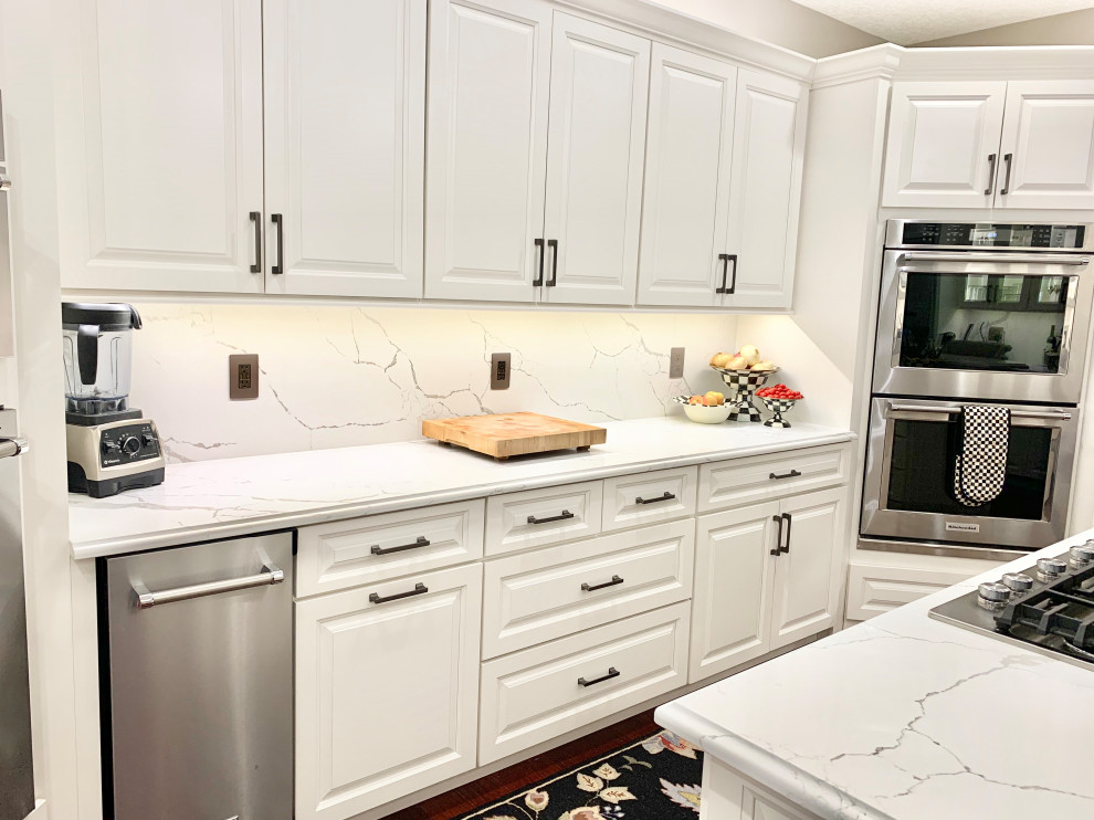 Photo of a large traditional u-shaped enclosed kitchen in Other with a belfast sink, raised-panel cabinets, white cabinets, engineered stone countertops, white splashback, stone slab splashback, stainless steel appliances, medium hardwood flooring, multiple islands and white worktops.