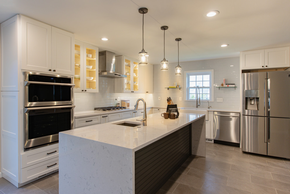 Photo of a large contemporary l-shaped kitchen/diner in DC Metro with a belfast sink, shaker cabinets, white cabinets, engineered stone countertops, white splashback, glass tiled splashback, stainless steel appliances, porcelain flooring, an island, grey floors and white worktops.