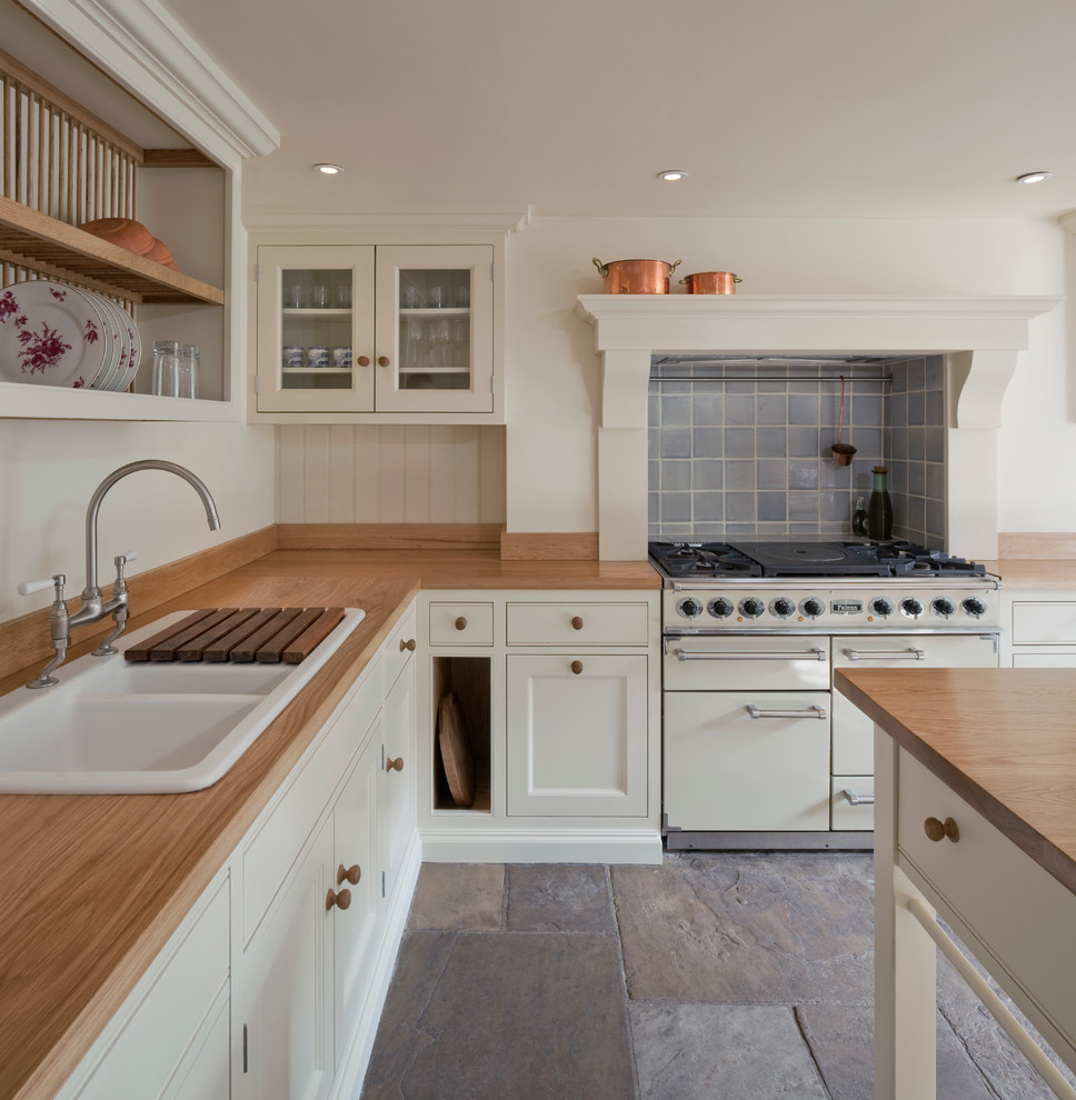 This is an example of a medium sized country kitchen in London with a belfast sink, shaker cabinets, beige cabinets, wood worktops, blue splashback, ceramic splashback, white appliances, slate flooring and an island.