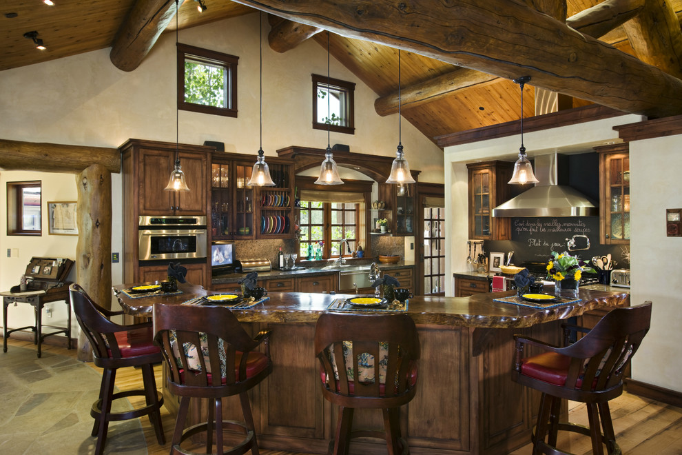 Cette image montre une grande cuisine américaine chalet en L et bois foncé avec un évier 2 bacs, un placard à porte vitrée, un plan de travail en bois, une crédence en dalle de pierre, un sol en bois brun et îlot.
