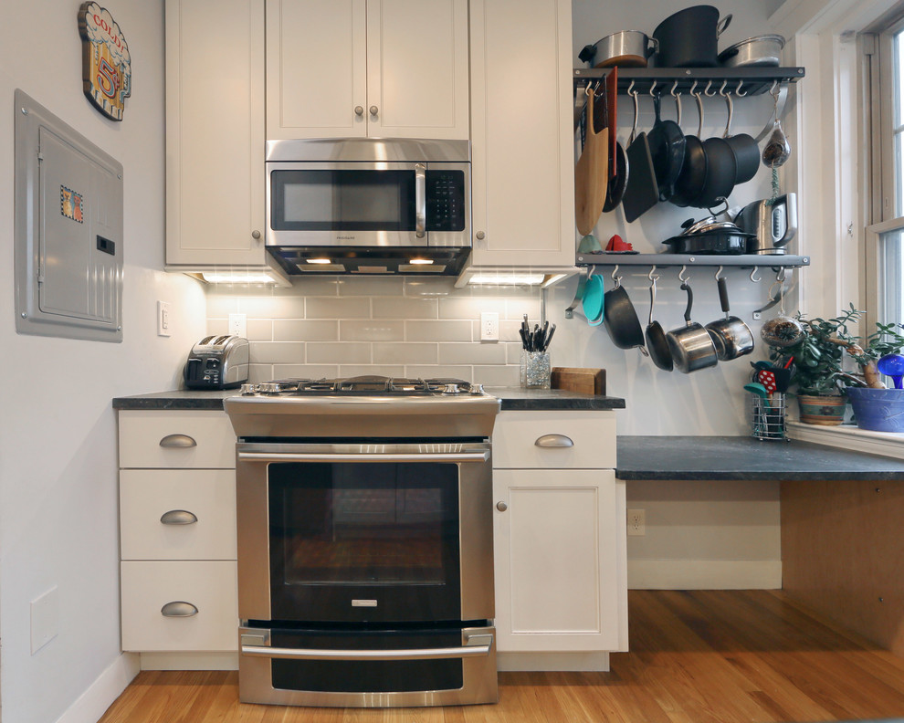 Example of a small transitional galley medium tone wood floor enclosed kitchen design in Boston with an undermount sink, shaker cabinets, white cabinets, granite countertops, gray backsplash, subway tile backsplash, stainless steel appliances and an island