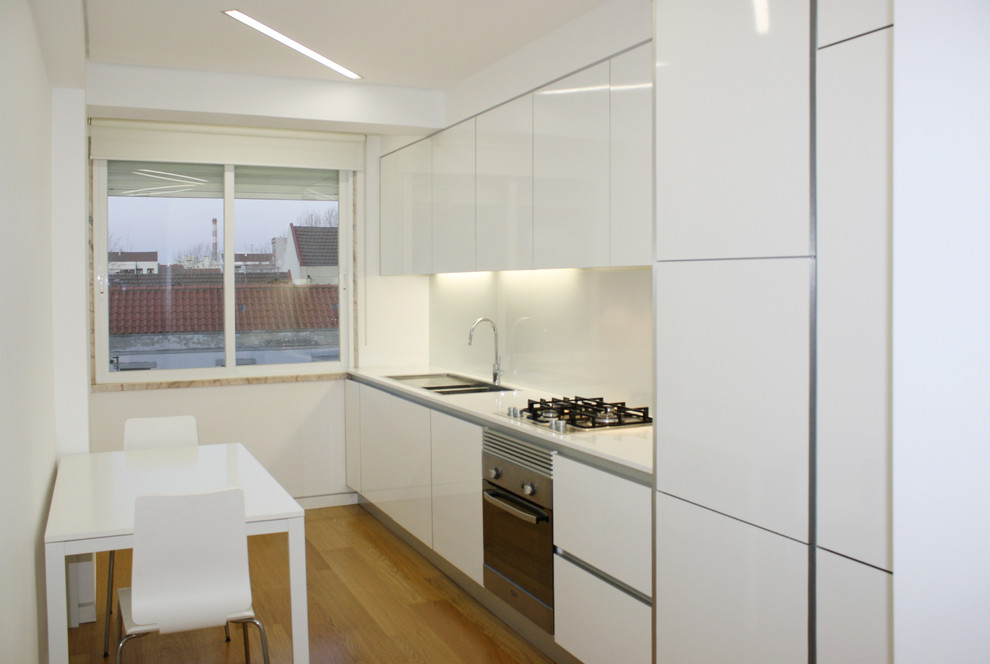 Cette image montre une cuisine minimaliste avec un évier encastré, un placard à porte plane, des portes de placard blanches, une crédence en feuille de verre et un électroménager en acier inoxydable.