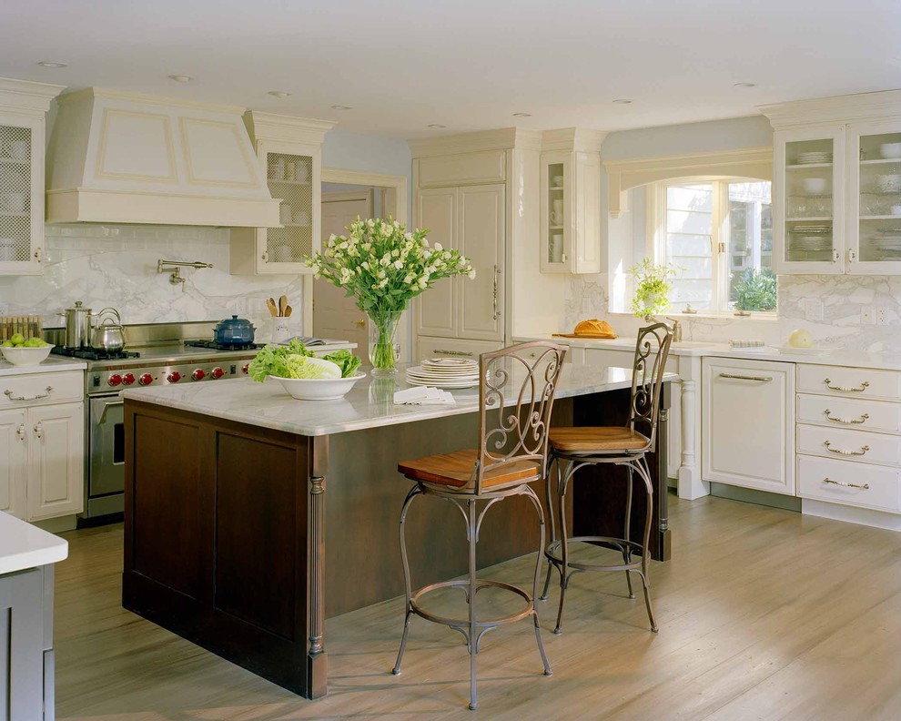 Modelo de cocina clásica de tamaño medio con armarios tipo vitrina, electrodomésticos con paneles, puertas de armario beige, encimera de mármol, salpicadero blanco, salpicadero de azulejos de piedra, suelo de madera clara, fregadero bajoencimera y barras de cocina