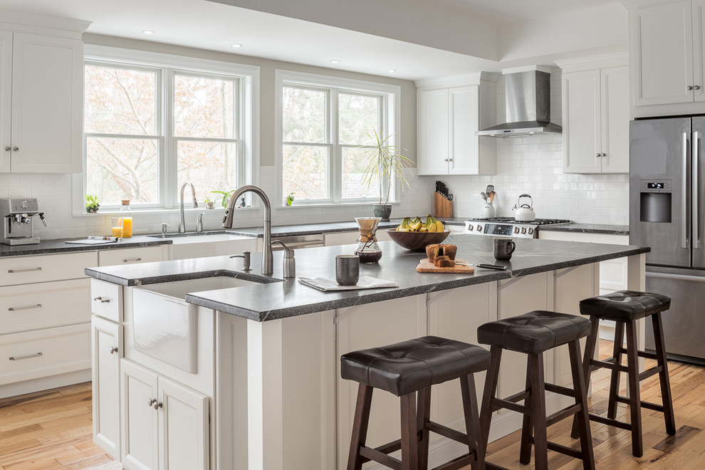 Klassische Küche in L-Form mit Landhausspüle, Schrankfronten mit vertiefter Füllung, weißen Schränken, Küchenrückwand in Weiß, Küchengeräten aus Edelstahl, braunem Holzboden, Kücheninsel, braunem Boden und grauer Arbeitsplatte in Portland Maine