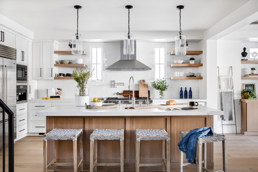 Inspiration for a large coastal u-shaped kitchen/diner in Orange County with a belfast sink, recessed-panel cabinets, white cabinets, engineered stone countertops, white splashback, metro tiled splashback, stainless steel appliances, light hardwood flooring, an island, beige floors and white worktops.