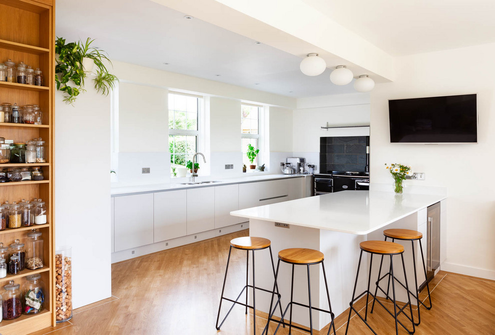 Contemporary u-shaped kitchen in Oxfordshire with a submerged sink, flat-panel cabinets, white cabinets, black appliances, medium hardwood flooring, a breakfast bar, brown floors and white worktops.