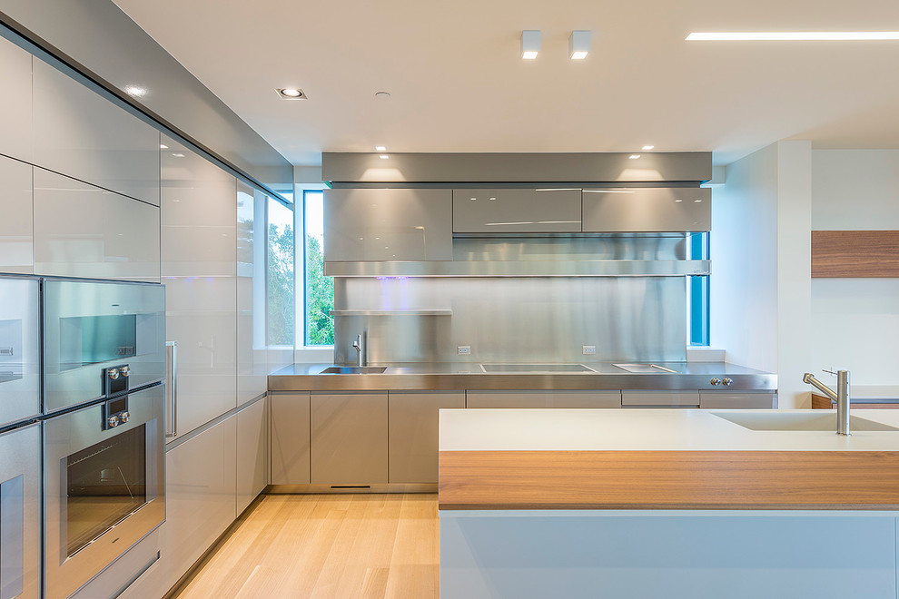 Bild på ett mellanstort funkis kök, med en integrerad diskho, släta luckor, grå skåp, bänkskiva i rostfritt stål, stänkskydd med metallisk yta, stänkskydd i metallkakel, rostfria vitvaror, ljust trägolv, flera köksöar och beiget golv