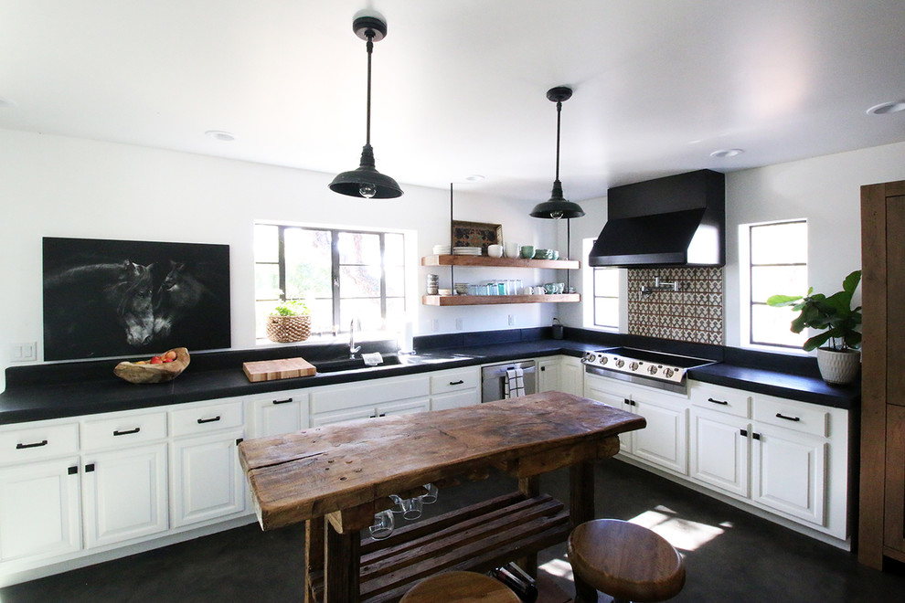 Пример оригинального дизайна: угловая кухня-гостиная в стиле кантри с монолитной мойкой, фасадами с выступающей филенкой, белыми фасадами, разноцветным фартуком, техникой из нержавеющей стали, островом и черным полом
