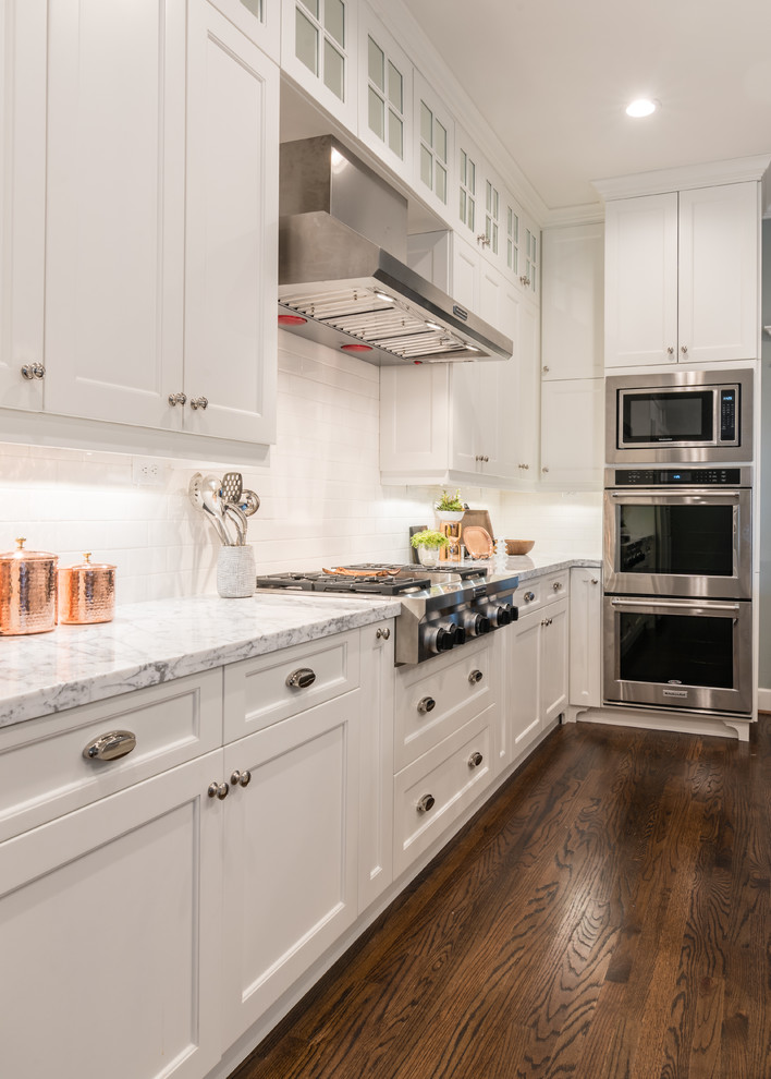 Large farmhouse l-shaped medium tone wood floor eat-in kitchen photo in Houston with a farmhouse sink, shaker cabinets, white cabinets, marble countertops, white backsplash, subway tile backsplash, stainless steel appliances and an island