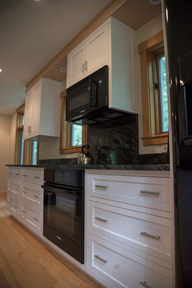 Mittelgroße Klassische Wohnküche in U-Form mit Doppelwaschbecken, flächenbündigen Schrankfronten, weißen Schränken, Granit-Arbeitsplatte, Küchenrückwand in Schwarz, schwarzen Elektrogeräten, hellem Holzboden und Kücheninsel in Boston