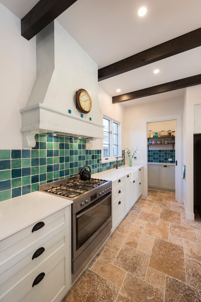 This is an example of a large mediterranean galley kitchen pantry in Melbourne with shaker cabinets, white cabinets, laminate countertops, multi-coloured splashback, terracotta splashback, an island, a belfast sink, stainless steel appliances and travertine flooring.