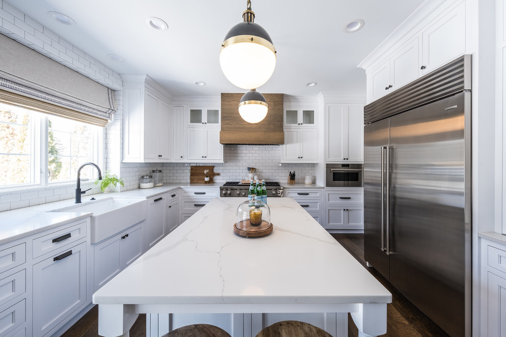 This is an example of a medium sized farmhouse u-shaped kitchen in Detroit with a belfast sink, recessed-panel cabinets, white cabinets, white splashback, metro tiled splashback, stainless steel appliances, dark hardwood flooring, an island, brown floors and white worktops.