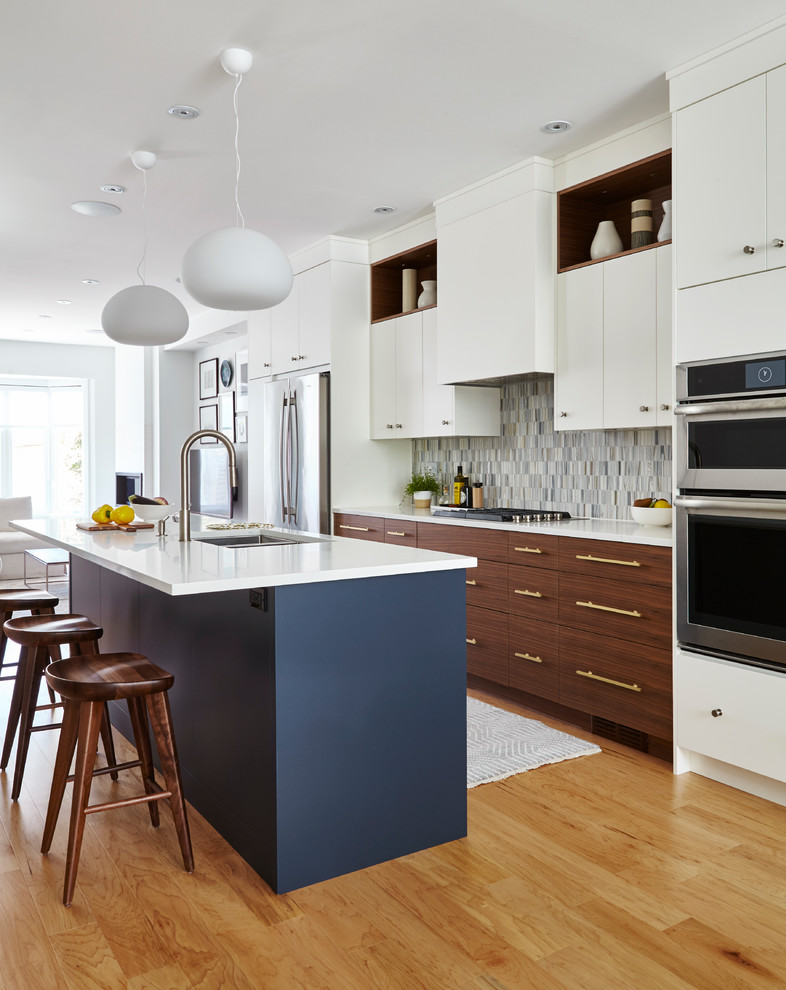 Inredning av ett modernt kök med öppen planlösning, med en undermonterad diskho, släta luckor, vita skåp, flerfärgad stänkskydd, rostfria vitvaror, mellanmörkt trägolv och en köksö