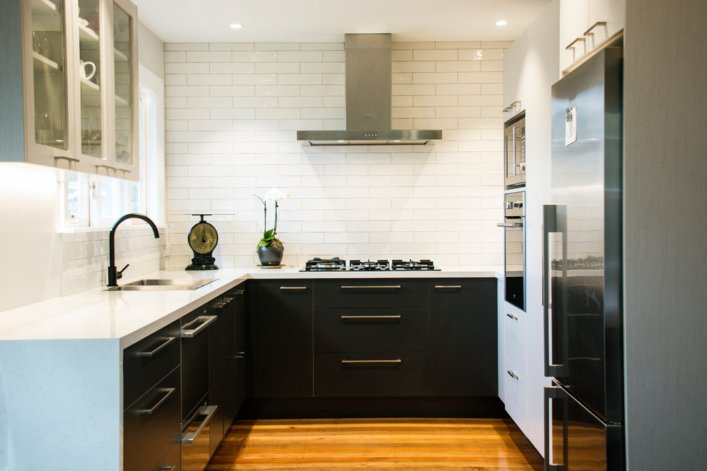 Bild på ett mellanstort funkis kök, med en dubbel diskho, svarta skåp, laminatbänkskiva, vitt stänkskydd, stänkskydd i tunnelbanekakel, rostfria vitvaror och mellanmörkt trägolv
