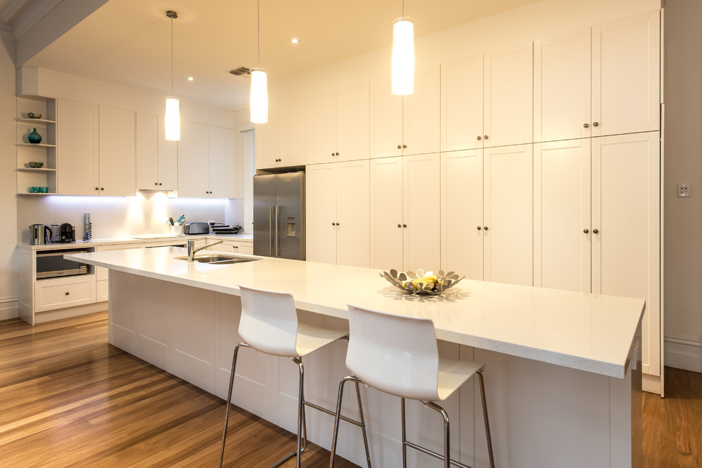 Klassische Küche in L-Form mit Doppelwaschbecken, Schrankfronten im Shaker-Stil, weißen Schränken, Küchengeräten aus Edelstahl, braunem Holzboden, Kücheninsel, braunem Boden und weißer Arbeitsplatte in Adelaide