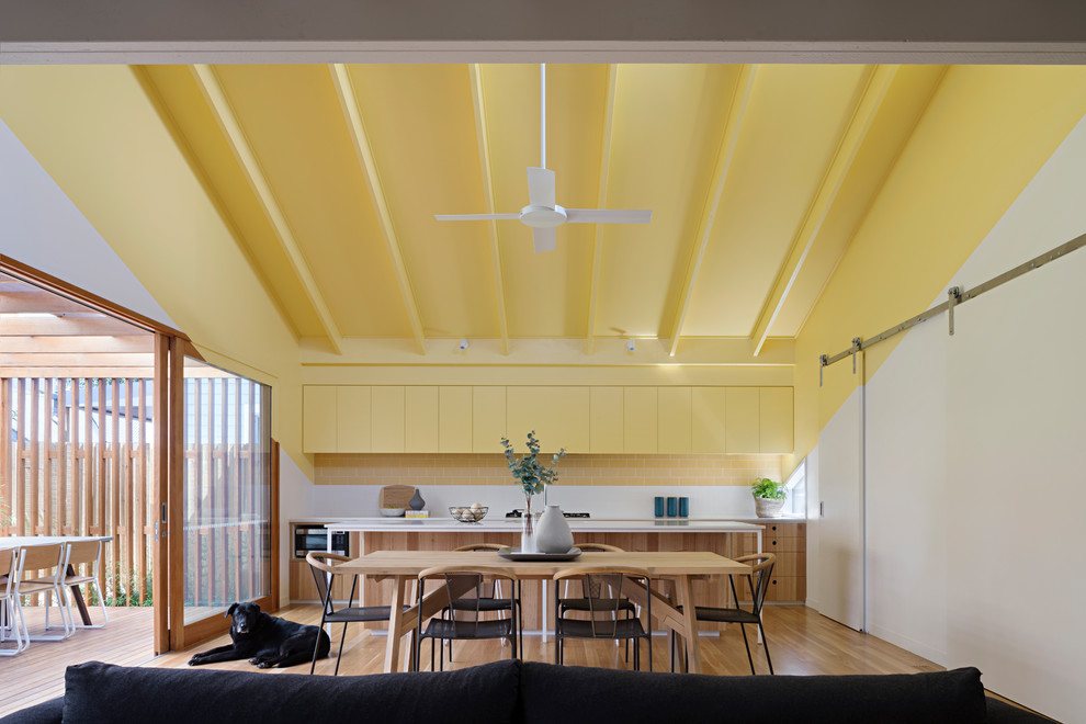 На фото: параллельная кухня-гостиная в современном стиле с плоскими фасадами, желтыми фасадами, желтым фартуком, фартуком из плитки кабанчик, паркетным полом среднего тона, островом и коричневым полом