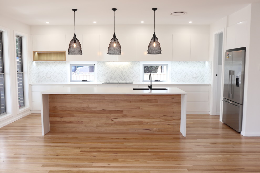 This is an example of a large modern galley kitchen in Wollongong with a double-bowl sink, flat-panel cabinets, white cabinets, engineered stone countertops, grey splashback, ceramic splashback, stainless steel appliances, light hardwood flooring, an island and white worktops.