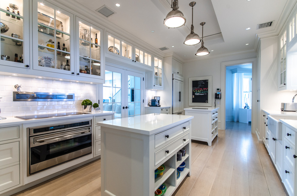 Inspiration for a large nautical galley kitchen in Miami with glass-front cabinets, white cabinets, engineered stone countertops, white splashback, metro tiled splashback, multiple islands, a belfast sink, stainless steel appliances and light hardwood flooring.