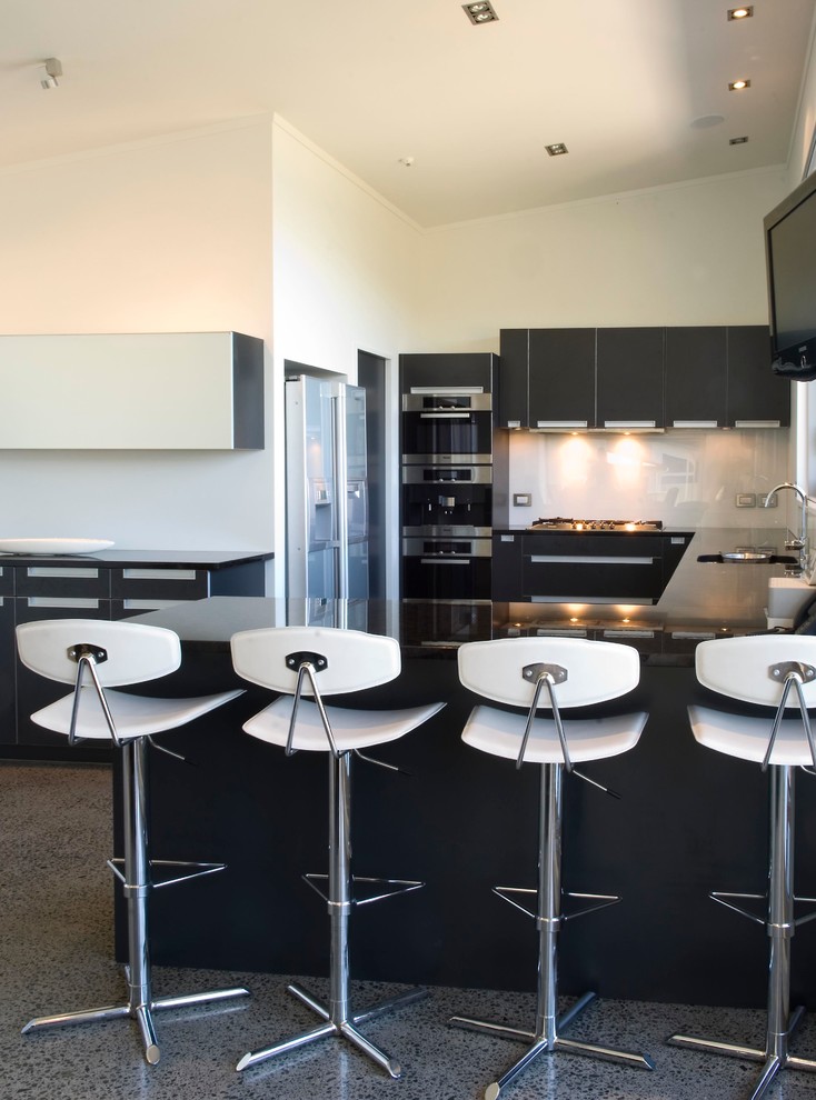 На фото: кухня в стиле модернизм с обеденным столом, одинарной мойкой, серыми фасадами, гранитной столешницей, белым фартуком, фартуком из стекла, черной техникой и бетонным полом без острова