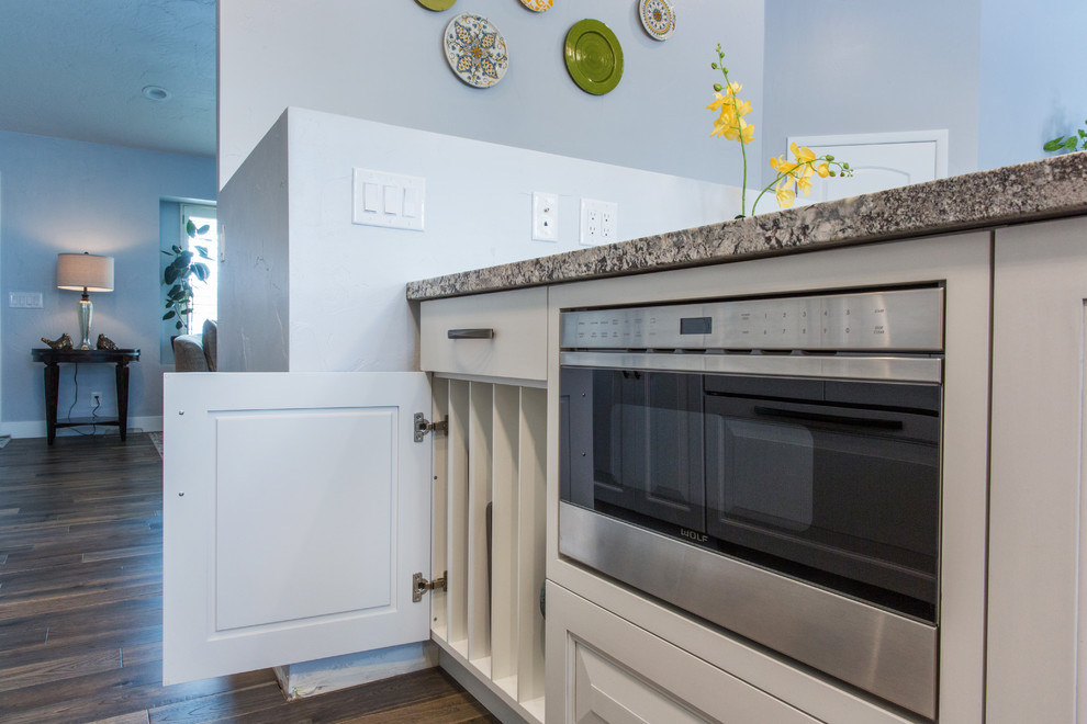 Example of a mid-sized transitional l-shaped medium tone wood floor eat-in kitchen design in Salt Lake City with an undermount sink, raised-panel cabinets, white cabinets, granite countertops, gray backsplash, glass tile backsplash, paneled appliances and a peninsula
