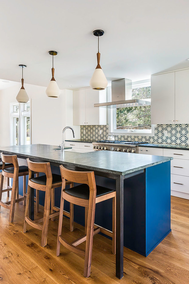 Modelo de cocina comedor actual con fregadero sobremueble, armarios con paneles lisos, puertas de armario blancas, electrodomésticos de acero inoxidable, suelo de madera en tonos medios, una isla, encimera de esteatita, salpicadero azul, salpicadero de azulejos de cemento, suelo marrón y encimeras grises