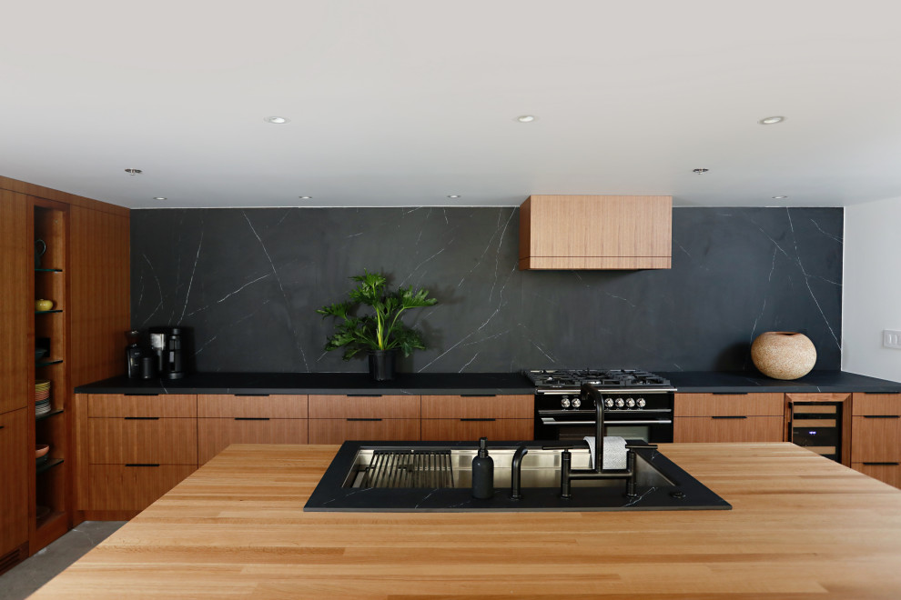 Modern inredning av ett svart svart l-kök, med en undermonterad diskho, släta luckor, skåp i mellenmörkt trä, svart stänkskydd, svarta vitvaror, betonggolv, en köksö och grått golv