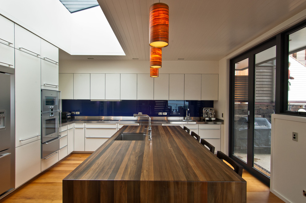 Moderne Wohnküche in L-Form mit Unterbauwaschbecken, weißen Schränken, Arbeitsplatte aus Holz, Küchenrückwand in Blau, Glasrückwand, Küchengeräten aus Edelstahl, braunem Holzboden und Kücheninsel in Wellington