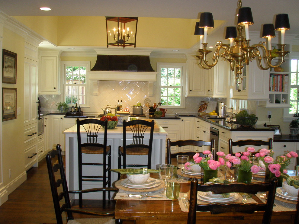На фото: п-образная кухня в классическом стиле с обеденным столом, фасадами с выступающей филенкой, белым фартуком, черно-белыми фасадами и барной стойкой