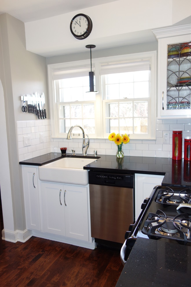 Small traditional l-shaped enclosed kitchen in Milwaukee with a belfast sink, shaker cabinets, white cabinets, engineered stone countertops, white splashback, ceramic splashback, stainless steel appliances, dark hardwood flooring and brown floors.