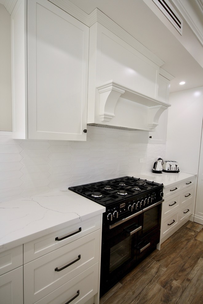 Example of a large classic l-shaped medium tone wood floor and brown floor eat-in kitchen design in Sydney with a farmhouse sink, shaker cabinets, white cabinets, quartz countertops, white backsplash, ceramic backsplash, black appliances, an island and white countertops