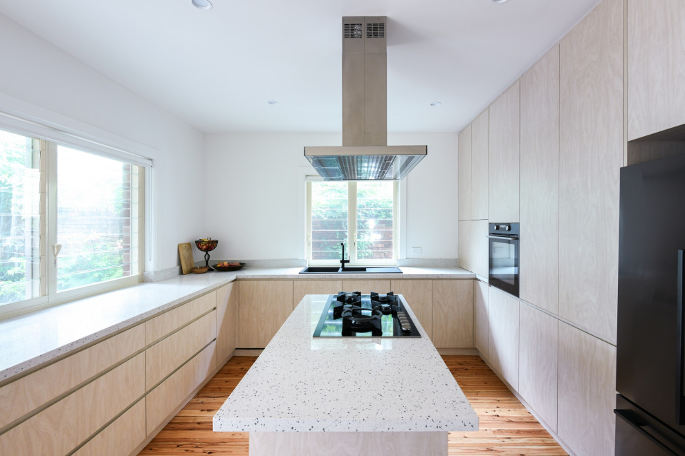 Inspiration for a large contemporary u-shaped enclosed kitchen in Sydney with a built-in sink, flat-panel cabinets, light wood cabinets, engineered stone countertops, beige splashback, engineered quartz splashback, black appliances, medium hardwood flooring, an island, brown floors and beige worktops.