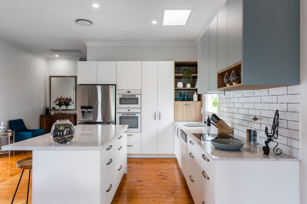 Mid-sized trendy l-shaped medium tone wood floor and brown floor open concept kitchen photo in Brisbane with an undermount sink, flat-panel cabinets, white cabinets, quartz countertops, white backsplash, ceramic backsplash, white appliances and an island