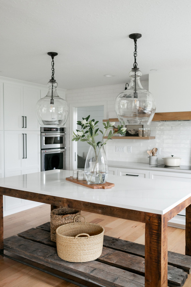 Design ideas for a medium sized country l-shaped kitchen/diner in Portland with a belfast sink, shaker cabinets, white cabinets, engineered stone countertops, white splashback, ceramic splashback, white appliances, light hardwood flooring, an island, brown floors and grey worktops.