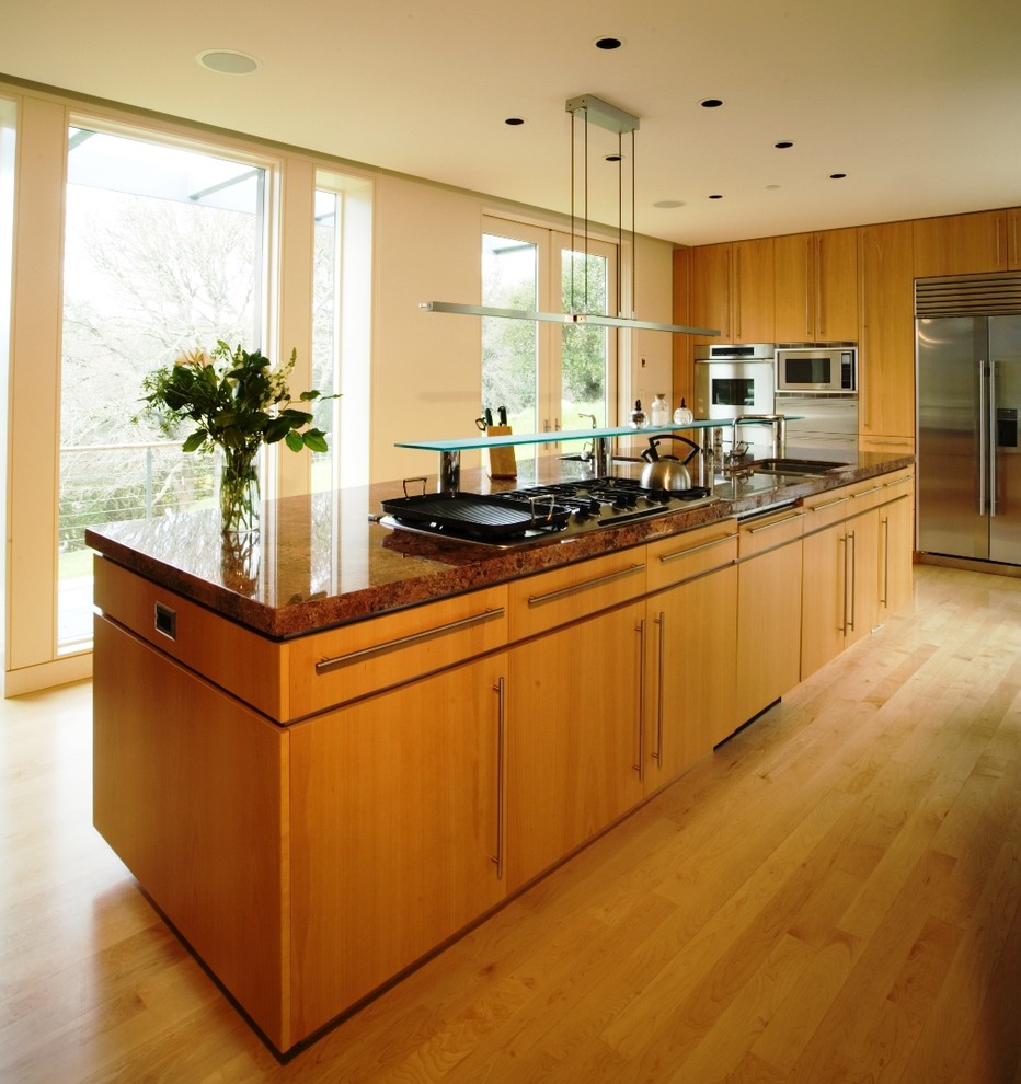 Cette image montre une grande cuisine design en bois brun et L fermée avec un électroménager en acier inoxydable, un placard à porte plane, un plan de travail en granite, parquet clair et îlot.