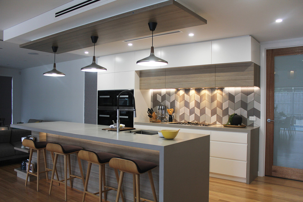 Photo of a medium sized contemporary galley open plan kitchen in Sydney with a submerged sink, flat-panel cabinets, white cabinets, engineered stone countertops, grey splashback, porcelain splashback, stainless steel appliances, medium hardwood flooring, an island and grey worktops.