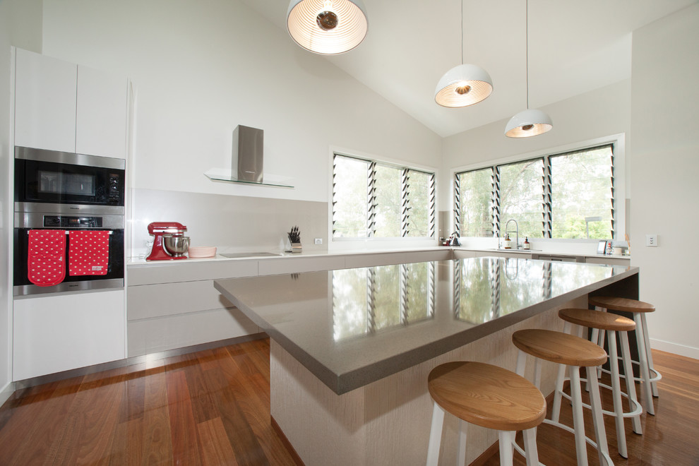 Moderne Wohnküche in L-Form mit Unterbauwaschbecken, flächenbündigen Schrankfronten, beigen Schränken, Quarzwerkstein-Arbeitsplatte, Küchenrückwand in Beige, Glasrückwand, Küchengeräten aus Edelstahl, braunem Holzboden und Kücheninsel in Brisbane