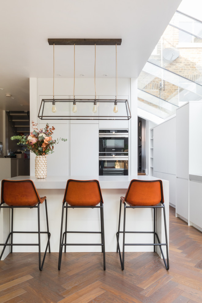 Moderne Wohnküche mit flächenbündigen Schrankfronten, weißen Schränken, braunem Holzboden und Kücheninsel in London