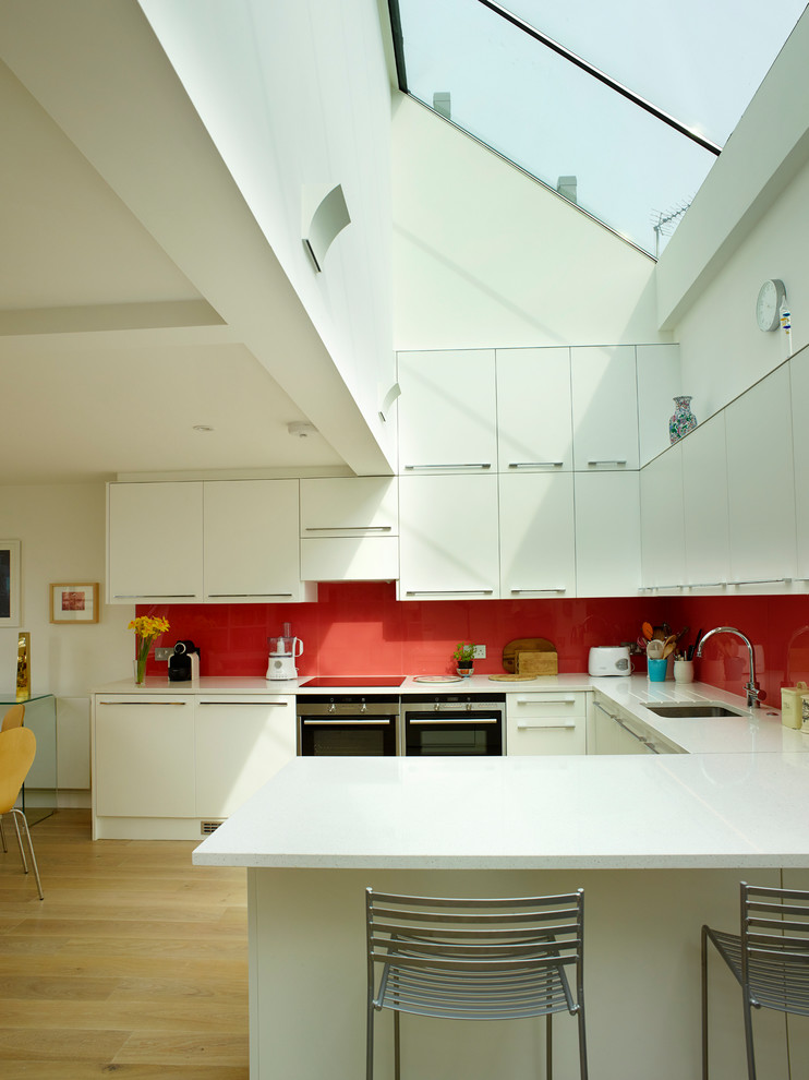 Inspiration for a contemporary u-shaped open plan kitchen in London with a submerged sink, flat-panel cabinets, white cabinets, red splashback, black appliances, light hardwood flooring and a breakfast bar.