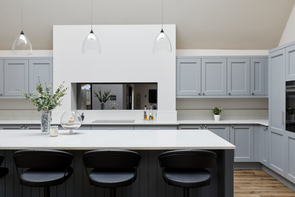 Large farmhouse u-shaped open plan kitchen in Kent with a belfast sink, shaker cabinets, grey cabinets, quartz worktops, mirror splashback, stainless steel appliances, medium hardwood flooring, an island, brown floors, white worktops and a vaulted ceiling.