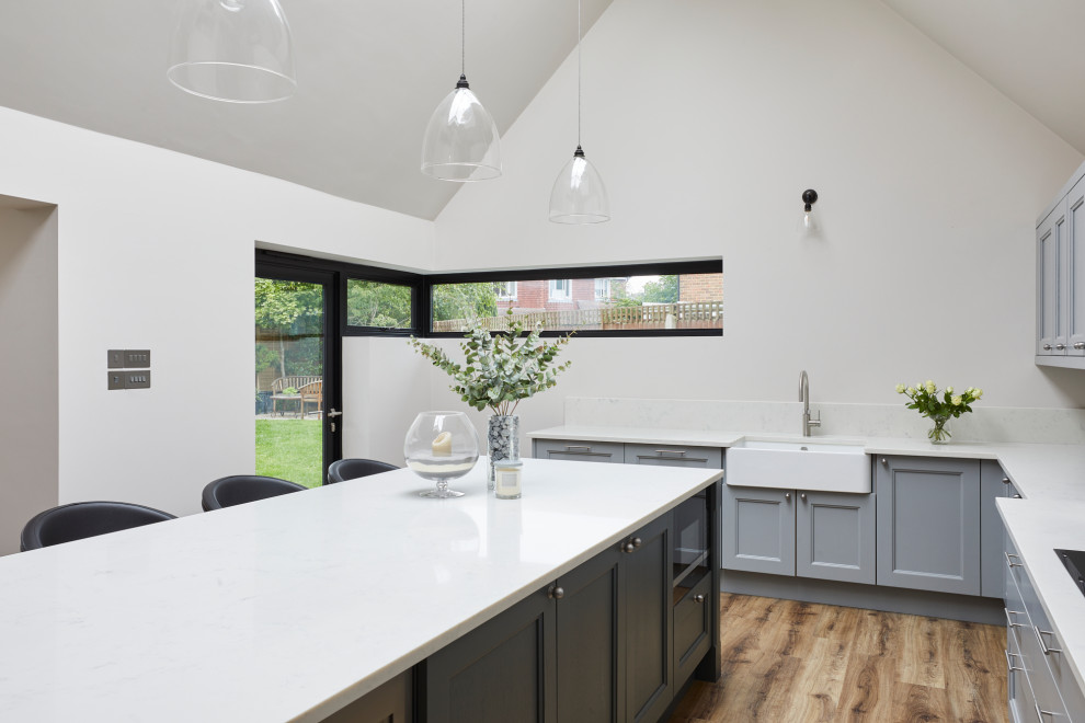 This is an example of a large farmhouse u-shaped open plan kitchen in Kent with a belfast sink, shaker cabinets, grey cabinets, quartz worktops, mirror splashback, stainless steel appliances, medium hardwood flooring, an island, brown floors, white worktops and a vaulted ceiling.