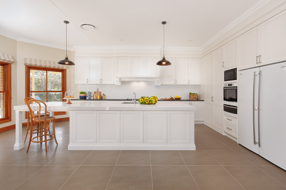 Design ideas for a large traditional single-wall open plan kitchen in Sydney with a submerged sink, shaker cabinets, white cabinets, engineered stone countertops, white splashback, ceramic splashback, white appliances, cement flooring, an island, beige floors and white worktops.