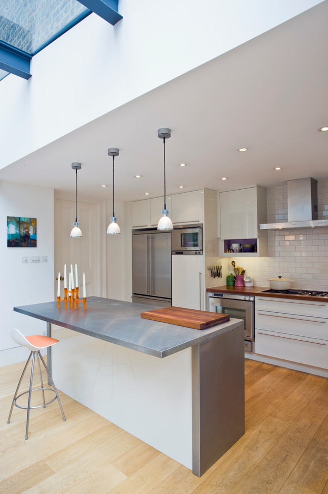 Photo of a contemporary galley kitchen in London with an island, flat-panel cabinets, stainless steel worktops, stainless steel appliances, light hardwood flooring, white cabinets, white splashback and metro tiled splashback.