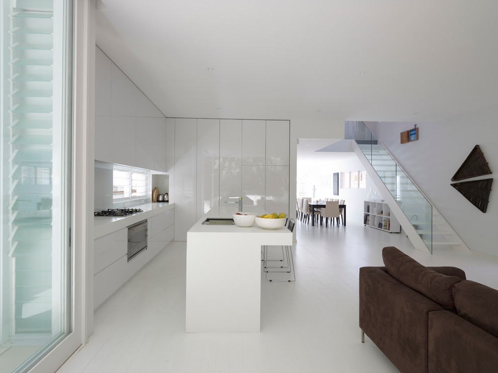На фото: угловая кухня-гостиная в современном стиле с накладной мойкой, плоскими фасадами, белыми фасадами, техникой под мебельный фасад, островом и белым полом