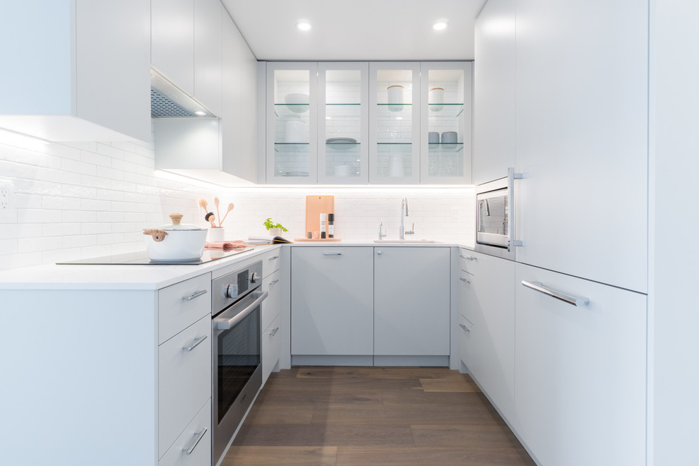 Modern inredning av ett litet vit vitt u-kök, med en enkel diskho, luckor med glaspanel, vita skåp, vitt stänkskydd, rostfria vitvaror, stänkskydd i tunnelbanekakel och mörkt trägolv